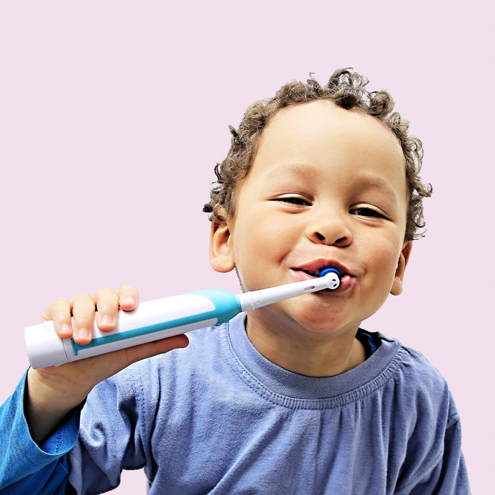 medium skin tone boy brushing teeth with an electric toothbrush and smiling, against pink background