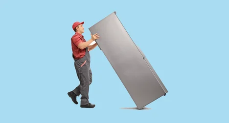 pale skinned man in work overalls pushing a large fridge on a plain background