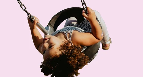 A medium skinned child on a tyre swing, mid-swing upside down, against a pink background.