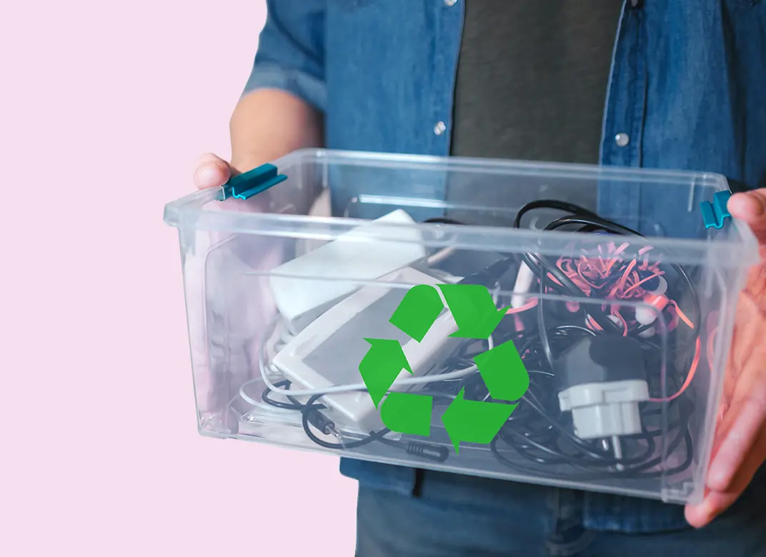 who-pays-weee--carrying-clear-box-1072x780-copy A light skinned man holding a see through plastic box full of electrical goods with a green recycle logo on the front against a pink background.