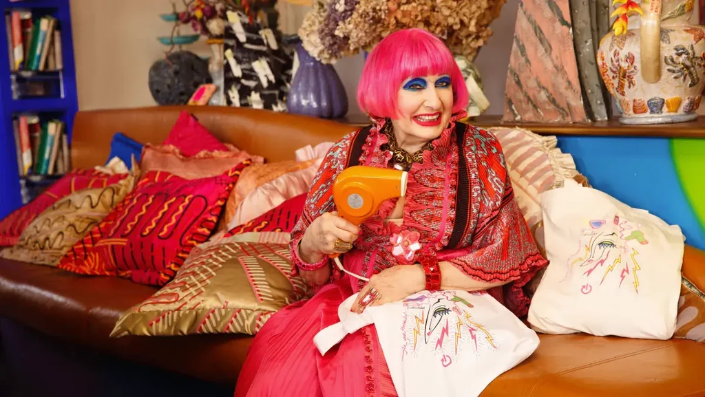 Light skinned fashion designer Zandra Rhodes wearing pink with pink hair holding electrical tote bag she designed and a hairdryer, sitting on a sofa with bright cushions